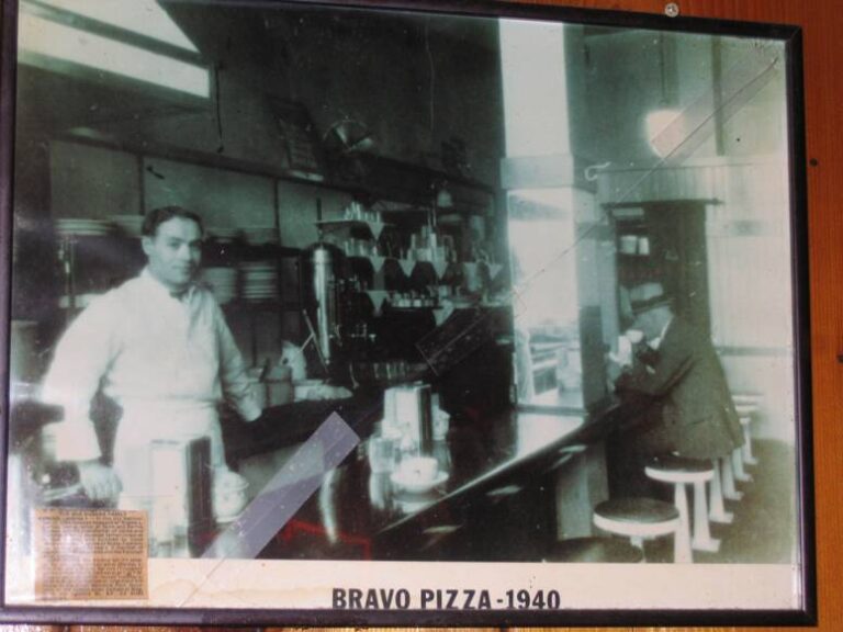 Framed 1940 photo of a man in a white apron behind the Bravo Pizza counter
